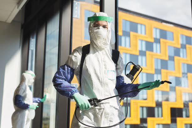 Waist up portrait of female worker wearing hazmat suit and holding disinfecting equipment looking at camera while standing in office, copy space